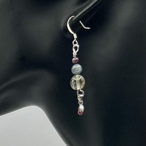 May include: Silver dangle earrings with a light gray, clear, and dark purple bead. The earrings have a silver hook closure.