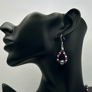 May include: Silver dangle earrings with a teardrop shape. The earrings are made with small pink beads and a single gray pearl.