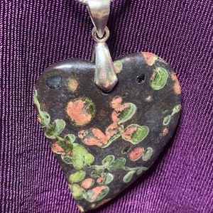 May include: A heart-shaped pendant made of dark brown stone with green and pink flecks. The pendant is attached to a silver chain.