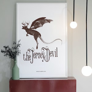 Könnte beinhalten: Ein gerahmter Druck einer braunen und weißen Illustration einer mythischen Kreatur mit Flügeln und einem langen Schwanz. Die Kreatur fliegt in der Luft und hat einen Hirschkopf. Der Text "the Jersey Devil" ist in einer dekorativen Schrift unter der Kreatur geschrieben. Der Text "SAGE PALMER 2022" ist in einer kleineren Schrift unter dem Text "the Jersey Devil" geschrieben.