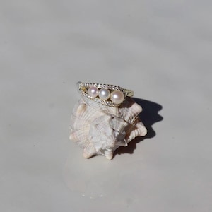 Può includere: Un anello dorato con tre perle di diverse tonalità, disposte in fila. La fascia è ornata da piccole gemme trasparenti. L'anello è esposto su una conchiglia bianca, creando un contrasto di trame e colori.