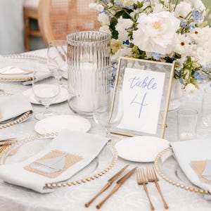 May include: Elegant table setting with clear glass plates, gold beaded rims, and white napkins featuring a sailboat design. A clear glass vase with a white candle sits next to a framed table number. A floral arrangement adds color.