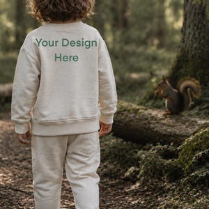 Könnte beinhalten: Ein cremefarbenes Set aus einem langärmeligen Sweatshirt und einer passenden Jogginghose. Auf der Rückseite des Sweatshirts befindet sich der dunkelgrüne Schriftzug "Your Design Here" in einer einfachen serifenlosen Schrift. Dazu weiße Sneaker.