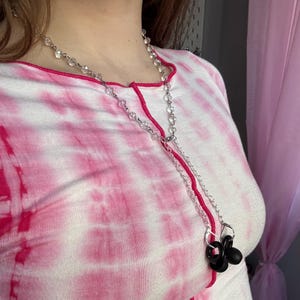 May include: A silver-toned necklace with clear, faceted beads and two black, circular pendants. The necklace is layered over a pink and white tie-dye shirt with a pink trim.