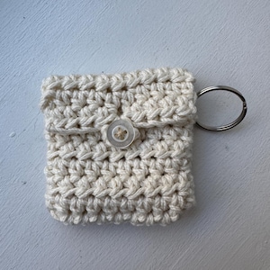 May include: A small, off-white crocheted pouch with a button closure and a silver key ring. The pouch is square-shaped and appears to be handmade. The button is a light tan color. The background is a light, textured surface.