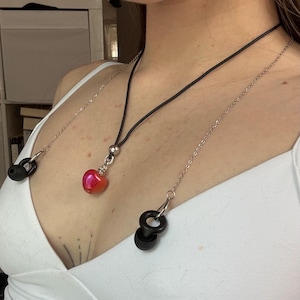 May include: A black cord necklace featuring a metallic silver bead and a glossy red heart-shaped pendant. Attached to the necklace are two silver-toned metal chains that connect to black silicone earbud holders, designed to secure wireless earbuds.