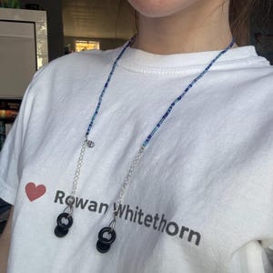 May include: A blue and white beaded glasses chain with black plastic earbud holders. The chain is worn by a person wearing a white t-shirt with the text "Rowan Whitethorn" printed on it.