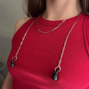 May include: A silver-toned metal chain necklace designed to hold wireless earbuds, featuring elongated oval links and small lobster claw clasps at each end that securely attach to a pair of black, rounded in-ear wireless earbud devices.