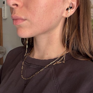 May include: A gold-toned metal chain with elongated rectangular links, designed to attach to an ear cuff or earring. The chain hangs down from the ear, draping loosely around the neck, creating a layered appearance with a thin silver-toned necklace.