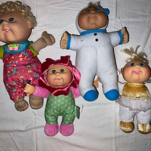 May include: Four Cabbage Patch Kids dolls in different outfits. One doll is wearing a pink and white outfit with a flower design, another is wearing a white astronaut suit with blue accents, another is wearing a green and white polka dot outfit with a pink flower on the head, and the last doll is wearing a gold and white outfit with a white angel wing.