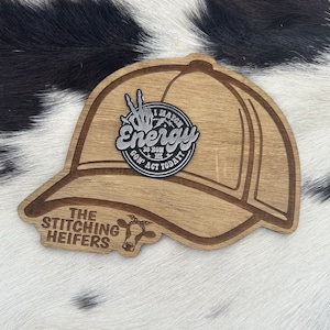May include: Wooden cutout of a baseball cap with a silver patch that says "I Match Energy So How Gon' Act Today?" and a cow head cutout below it that says "The Stitching Heifers".