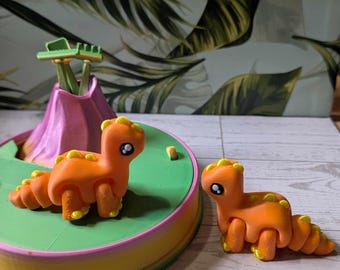 May include: Two orange and yellow articulated dinosaur toys on a green, pink, and yellow base. The dinosaurs have large eyes and are posed in a standing position.
