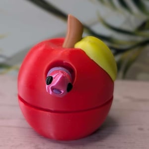 May include: A red apple shaped container with a pink worm poking out of the top. The apple is split in half and the top half is slightly open.