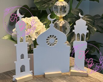 May include: Three white wooden cutout mosque decorations with intricate designs. The mosque on the left has a tall minaret, the middle mosque has a dome, and the mosque on the right has a smaller minaret. Each mosque is on a small wooden base.