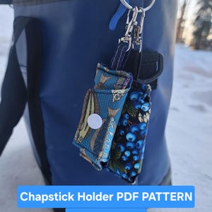 Puede incluir: Dos fundas para lápiz labial con diferentes diseños, sujetas a un llavero. Una funda tiene un diseño de peces azules, la otra un diseño de bayas azules. El texto "Chapstick Holder PDF PATTERN" y "Digital download only" es visible.
