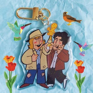 May include: A clear acrylic keyring with a cartoon image of two men holding a bottle. The keyring has a gold-coloured clasp. Decorative elements include a hummingbird, a robin, and tulips. The background is light blue.