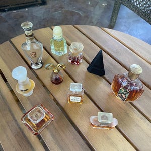 May include: A selection of miniature perfume bottles in diverse shapes and colours, including clear, amber, and red glass. Some bottles have decorative stoppers, and one is labelled "Chanel". The bottles are arranged on a wooden surface.
