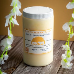 May include: A white jar of grass-fed beef soft tallow, labeled "GRASS FED BEEF SOFT TALLOW 100% Dry Rendered Back Trim Fat". The jar is on a wooden surface with white flowers in the background.