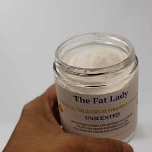 May include: A clear glass jar of The Fat Lady unscented whipped tallow cream. The label includes text about the ingredients: unfiltered honey and bovine colostrum. The cream is white and appears to be a skincare product.