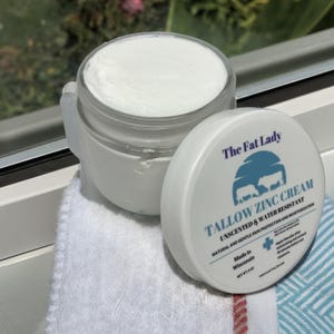 May include: A white jar of The Fat Lady Tallow Zinc Cream, unscented and water resistant. The jar is labeled with the product name, ingredients, and instructions for use. The jar is sitting on a white towel.