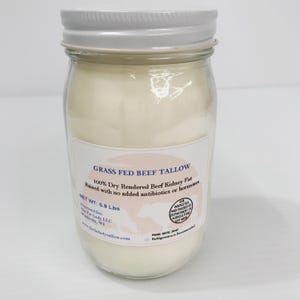 May include: A clear glass jar filled with off-white beef tallow, topped with a silver metal lid. The label reads: GRASS FED BEEF TALLOW, 100% Dry Rendered Beef Kidney Fat, Raised with no added antibiotics or hormones, NET WT: 0.9 Lbs.