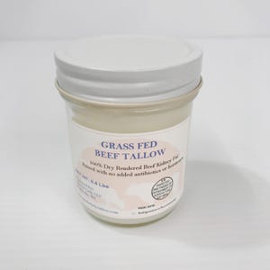 May include: A clear glass jar with a white metal lid containing off-white beef tallow. The label reads Grass Fed Beef Tallow, 100% Dry Rendered Beef Kidney Fat, Raised with no added antibiotics or hormones, and Net Wt: 0.4 Lbs.