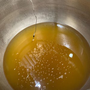 May include: A large stainless steel pot filled with a golden yellow liquid. A thermometer is submerged in the liquid.