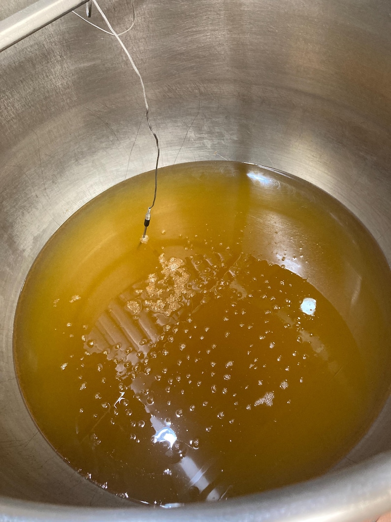 May include: A large stainless steel pot filled with a golden yellow liquid. A thermometer is submerged in the liquid.