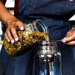 Puede incluir: Una persona vierte un l&iacute;quido de un frasco de vidrio con un colador en un frasco de vidrio transparente. El frasco contiene una mezcla de ingredientes marrones y amarillos.