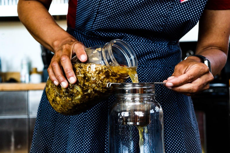 Puede incluir: Una persona vierte un l&iacute;quido de un frasco de vidrio con un colador en un frasco de vidrio transparente. El frasco contiene una mezcla de ingredientes marrones y amarillos.