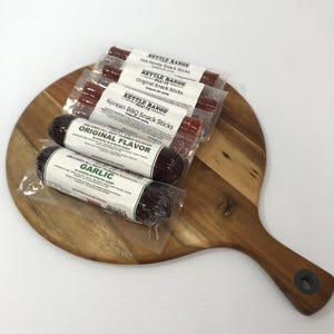 Può includere: Una collezione di snack stick e salumi estivi Kettle Range Meat Co. in confezioni di plastica trasparente, disposti su un tagliere rotondo in legno. Le etichette riportano testi tra cui i gusti Hot Honey, Original, Korean BBQ e Garlic.