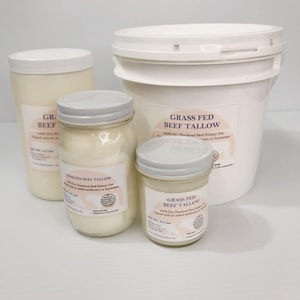 May include: A collection of four containers of grass fed beef tallow, including one large white plastic bucket and three glass jars of varying sizes, all featuring white lids and labels with the text Grass Fed Beef Tallow and net weights.