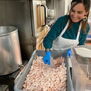 May include: A person wearing blue gloves scoops white and pink meat from a large gray container. The container is filled with small pieces of meat.