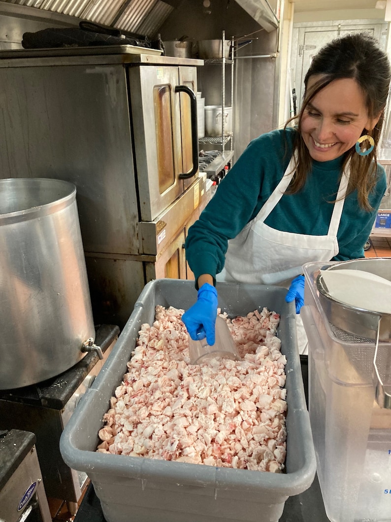 May include: A person wearing blue gloves scoops white and pink meat from a large gray container. The container is filled with small pieces of meat.