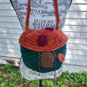 May include: A crocheted handbag with a mushroom design. The bag is made with a variety of colors, including green, brown, and orange. The bag has a long strap and a small pocket on the front.