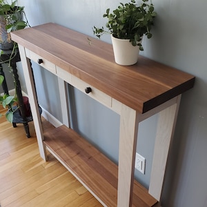 May include: A white console table with a walnut wood top and three drawers. The table has a shelf underneath the top. The table is standing on a wood floor.