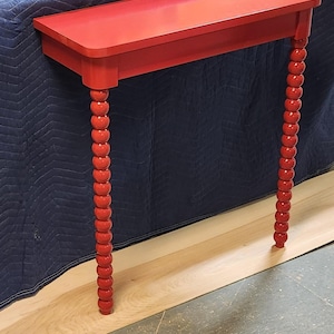 May include: A vibrant red console table with a rectangular top and two decorative, beaded legs. The table is made of wood and has a glossy finish. The legs are a unique design element, adding visual interest to the piece.