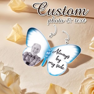 Op de afbeelding: Een vlindervormig aandenken met een foto van een lachende persoon en de tekst "Always by my side". De vlinder heeft blauwe en witte vleugels, versierd met blauwe edelstenen. De tekst "Custom photo & text" staat bovenaan.