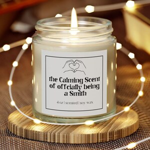 Könnte beinhalten: Ein Glas-Kerzengefäß mit einem Holzdeckel. Die Kerze ist angezündet und das Etikett lautet "The Calming Scent of Officially Being a Smith" mit einem Herzsymbol. Die Kerze ist eine 4 oz duftende Sojawachskerze.