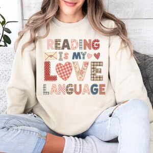 May include: Cream-colored sweatshirt with the text "READING IS MY LOVE LANGUAGE" in a patchwork design. The text includes hearts, an envelope, and floral patterns. The sweatshirt is a casual, comfortable style.