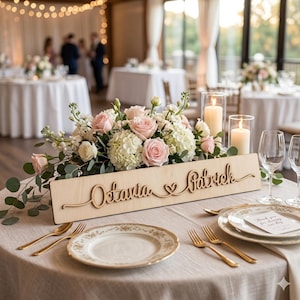 May include: A wedding reception table setting with a floral centerpiece of pink roses and white hydrangeas. A wooden sign with the names "Octavia & Patrick" is in front of the flowers. Gold-colored cutlery and plates are set for each guest.