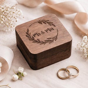 May include: A wooden ring box with the words "Mr & Mrs" engraved on the lid, surrounded by a laurel wreath design. Two gold wedding rings are next to the box. The box and rings are on a light-colored surface with floral accents.