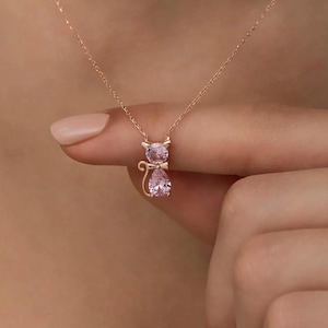 Puede incluir: Collar con colgante de gato en oro rosa con piedras preciosas rosas. El colgante de gato presenta una cabeza de piedra preciosa redonda y un cuerpo de piedra preciosa en forma de pera. El collar es delicado y femenino, perfecto para los amantes de los gatos. El collar lo sostiene una persona.