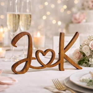 May include: Wooden initials "J & K" with a heart-shaped detail, ideal for wedding decor. The letters are in a warm brown tone, set on a white tablecloth with champagne flutes and candles in the background.