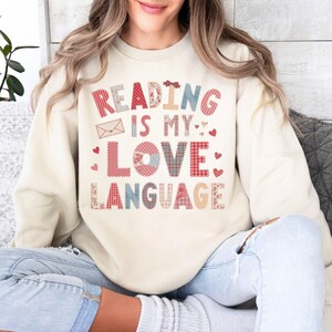 May include: Cream-colored sweatshirt with the text "READING IS MY LOVE LANGUAGE" in a patchwork design. The text includes hearts and an envelope graphic. The sweatshirt is a casual style.