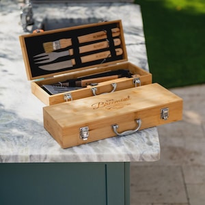 May include: A bamboo barbecue tool set in a wooden case. The case is open, revealing a black interior with various tools, including a brush, fork, and knife. The closed case has the text "The Breemont" on the lid. The set is on a marble surface.