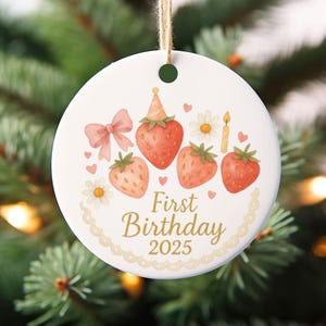May include: A white ceramic ornament with a watercolor design featuring strawberries, a bow, and the text "First Birthday 2025." The ornament is round and has a string for hanging. The design includes a birthday candle and small flowers.
