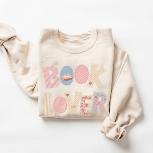 May include: Cream-colored sweatshirt with the words "BOOK LOVER" appliqued in various patterned fabrics. The design includes a stack of books and hearts, perfect for book enthusiasts.