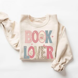 May include: Cream-colored sweatshirt with the words "BOOK LOVER" in a patchwork design. The letters are in pink, blue, and patterned fabrics, with a stack of books in an oval and small hearts. A bow is on the letter "R".