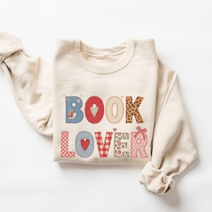 May include: A cream-coloured sweatshirt featuring the words "BOOK LOVER" appliquéd across the front. The letters are in various patterns and colours, including blue, red, leopard print, and pink. The sweatshirt has long sleeves.